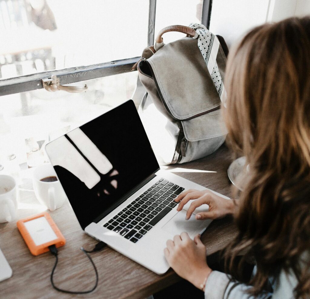 A young woman works remotely at a café, using her laptop and external hard drive.